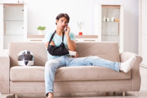 Motorcycle Accident Lawyer in Santa Barbara | Free Consultations 24/7 A motorcycle accident victim with his arm in a sling and foot in a cast sitting on a couch beside his helmet and calling an experienced motorcycle accident lawyer in Santa Barbara.